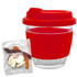 A glass coffee cup with a red silicone lid and sleeve, accompanied by a packet of mixed lollies in various colours.