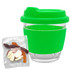 A clear glass coffee cup with a bright green silicone lid and sleeve, accompanied by a pack of mixed lollies.