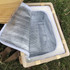 A square wicker picnic cooler basket with a wooden lid and a silver insulated interior, resting on green grass.