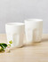 A set of two white latte cups with a textured design, placed on a wooden surface alongside a small flower.
