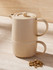 A beige tea set with a teapot and cup stacked together, accompanied by dried flowers, on a light stone surface.