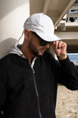 A man wearing a white cap and sunglasses stands under a structure, dressed in a black jacket with a hood.