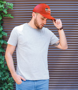 A man wearing a grey t-shirt and a red cap stands against a textured background with greenery. The cap features a logo.