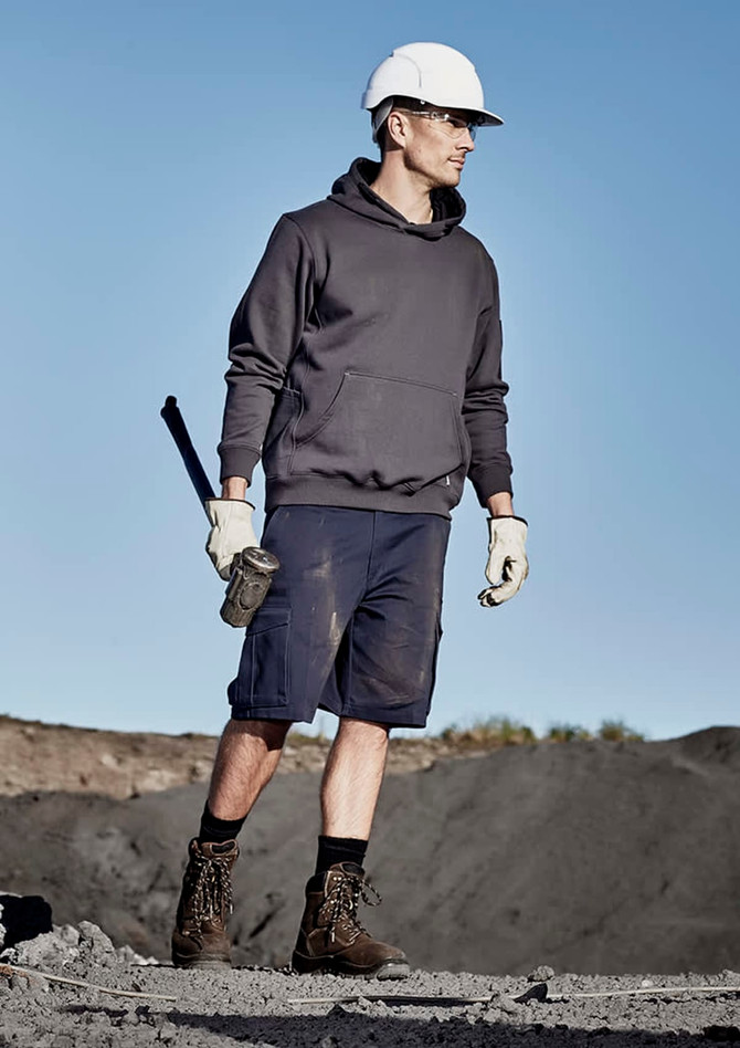 A man in a grey hoodie and navy cargo shorts stands outdoors, wearing a hard hat and gloves, holding a tool.