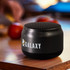 Auris Bluetooth speaker in black, featuring a textured top and a visible logo, placed on a wooden surface near apples.