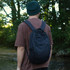 A black daypack worn on the back of a person in a forested setting, with green foliage in the background.