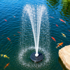 White floating fountain creating a tall vertical water spray in a clear blue pond with colorful koi fish (red, orange, and yellow) swimming below, demonstrating the fountain's aesthetic appeal in an aquatic garden setting.