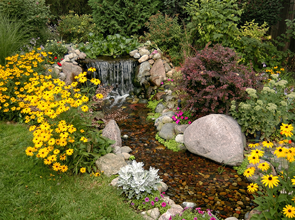 Backyard pondless waterfall flowing over rocks through a lush garden with yellow black-eyed susans and colorful flowering plants