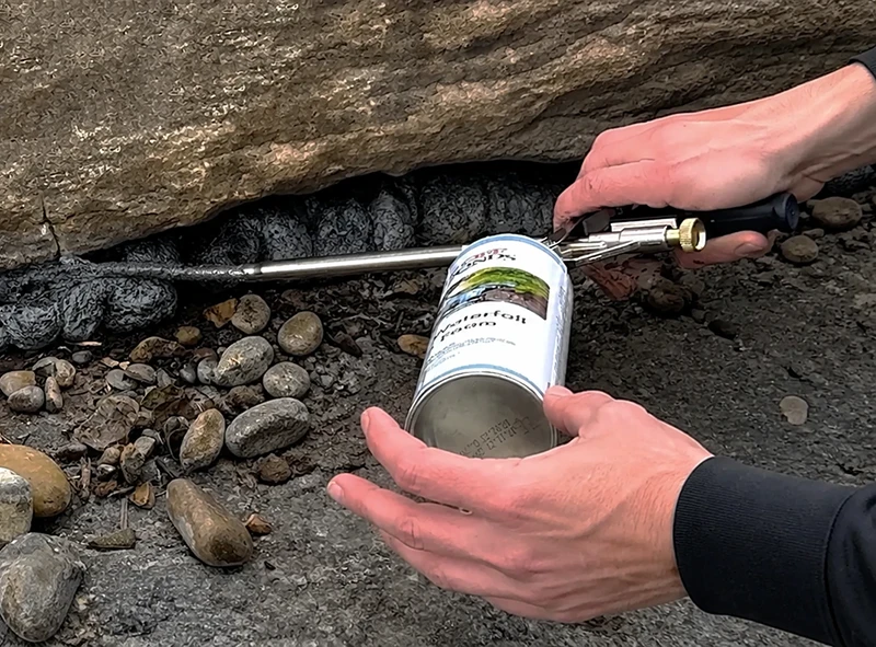 Applying black waterfall foam with a dispensing gun between rocks to seal gaps in a pond waterfall feature.
