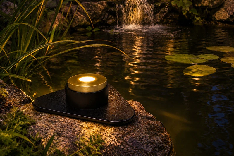 Pond puck light illuminating a rock ledge beside a koi pond at night, casting a warm glow across the surrounding water and landscape.