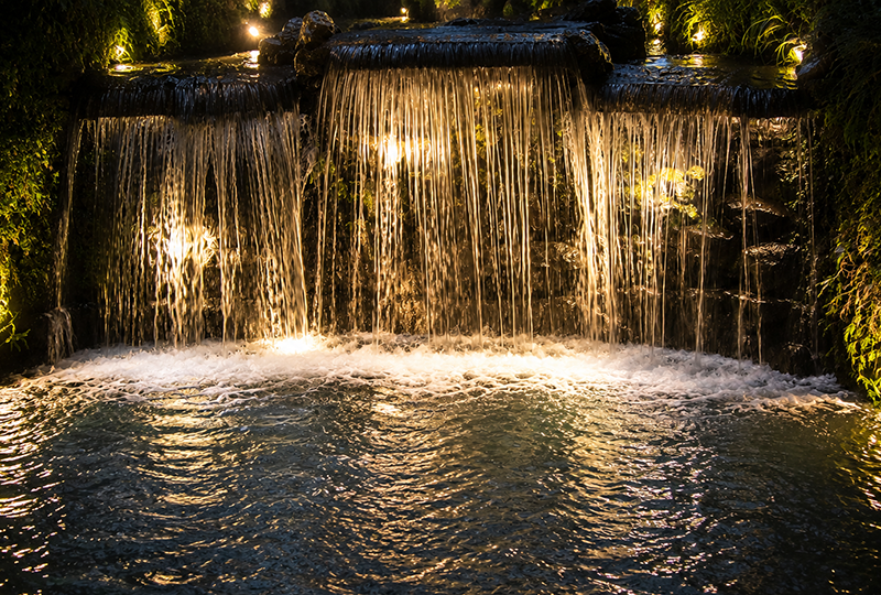 Illuminated outdoor waterfall spillway at night, with dramatic sheet water flow into pond, professional landscape lighting, and surrounding greenery