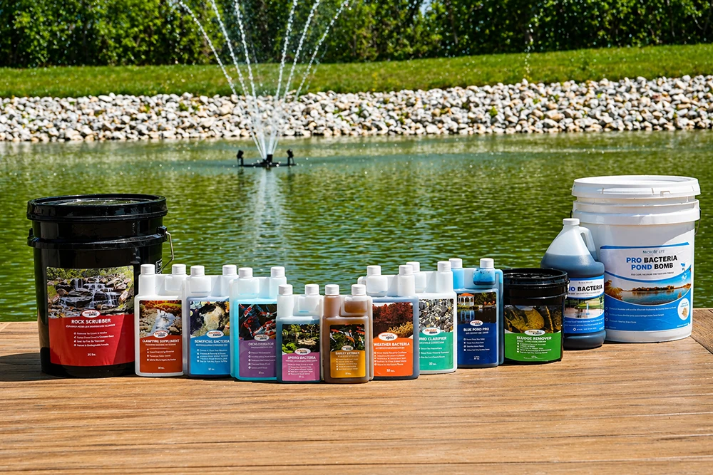 Lineup of Half Off Ponds water treatments and bacteria products displayed on a dock beside a pond with floating fountain