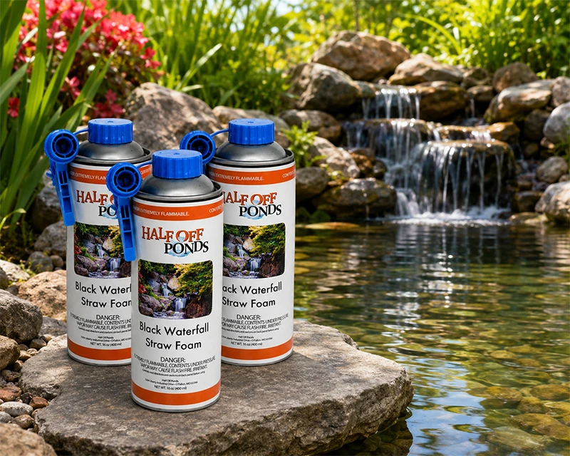 Black Waterfall Straw Foam sealant cans by Half Off Ponds displayed on a natural stone ledge beside a backyard pond waterfall with lush green and red foliage in the background