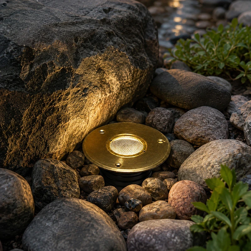 Round brass in-ground well light set among river rock, washing warm light up the face of a large boulder beside a stream.