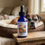 Bottle of Recovery For Pets flower essence on a wooden bench beside a senior boxer resting in a round dog bed in the recovery window after a vet visit.