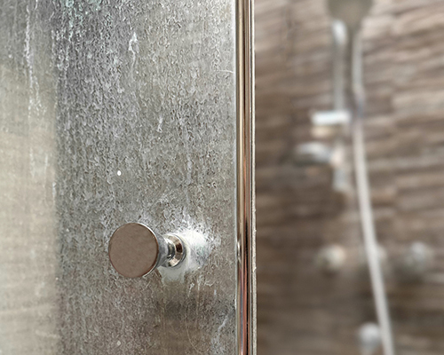 Close-up of a glass shower door affected by limescale partially open, with a chrome handle