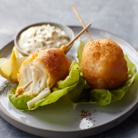 Two Alaska halibut corn dogs on a bed of lettuce with tartar sauce & lemon slices.