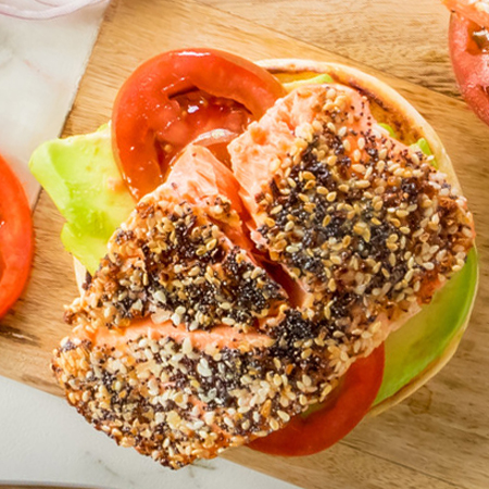 A spiced salmon fillet portion atop a toasted bagel with avocados and toppings