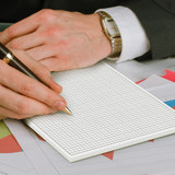 A hand holding a pen writes on a blank, gridded A4 notepad with a textured surface. The background features colourful paper.