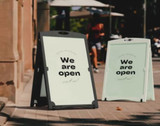Two premium plastic water-fillable corflute A-frames in black and white, standing on a footpath with people in the background.