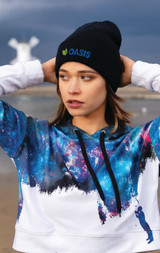 A model wearing a black beanie with a logo and a galaxy-print sweatshirt, set against a coastal background.