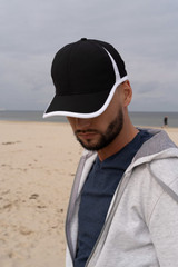 A black cap with white trim, worn by a person on a beach, with a cloudy sky in the background.
