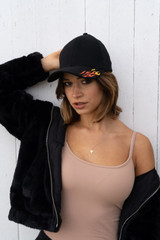 A black cap with flame graphics is worn by a model in a beige top and black jacket, posed against a white background.