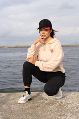 A woman squats by the water, wearing a beige hoodie and black pants, complemented by a black cap with red accents.