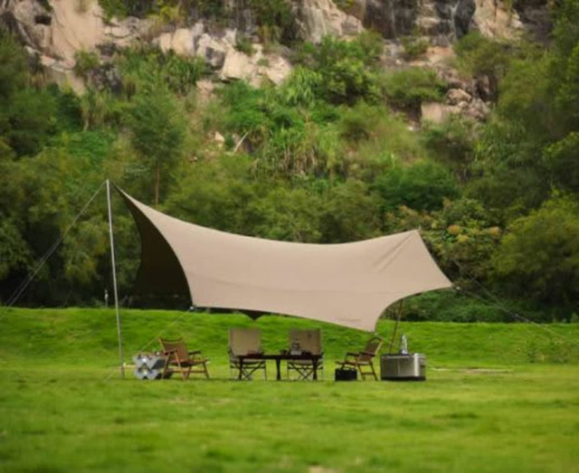 A tan camping canopy with poles, set up in a grassy area surrounded by greenery and rocky terrain.