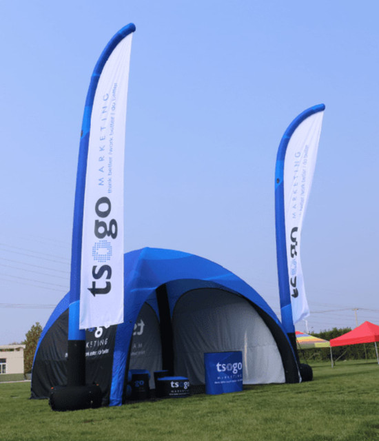 A blue and black inflatable tent with two tall white flags, featuring a logo, set on a grassy field.