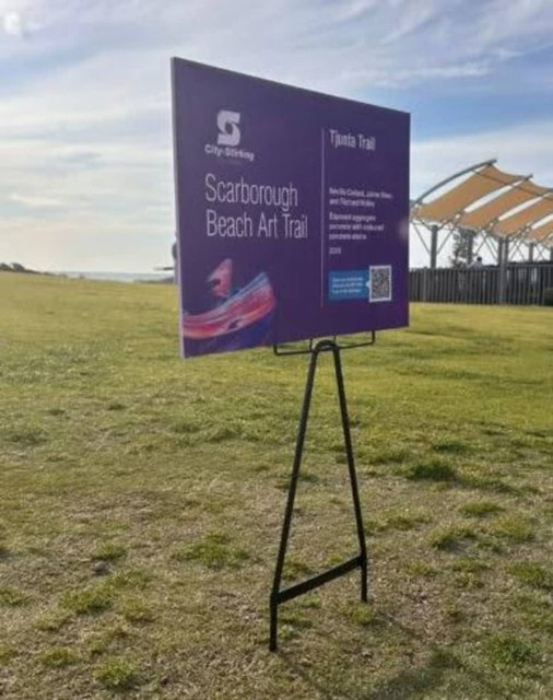 A promotional display featuring a purple sign with information about the Scarborough Beach Art Trail, on a black wire stand.