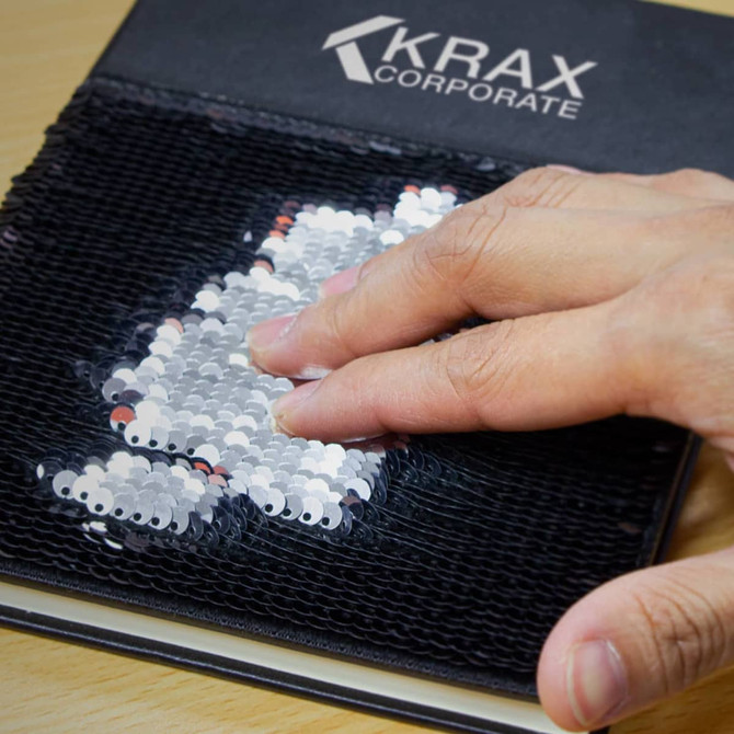 A hand glides over a black sequin notebook, revealing shifting silver and white sequins beneath. It has a logo on the cover.