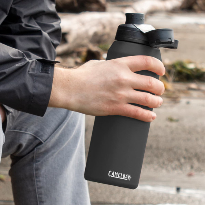 A drink bottle in matte black with a screw-top lid, held by a person sitting outdoors. It features a logo.