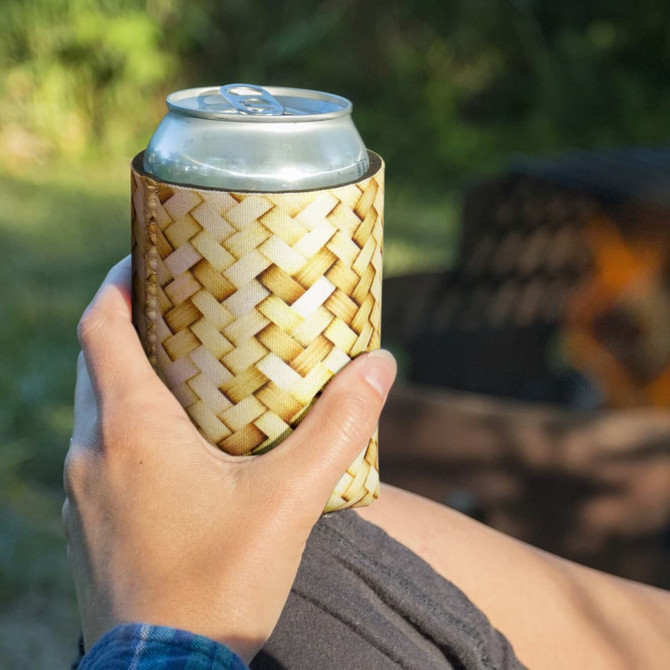A drink bottle cooler with a woven design, held by a hand outdoors, featuring natural tones and a logo.