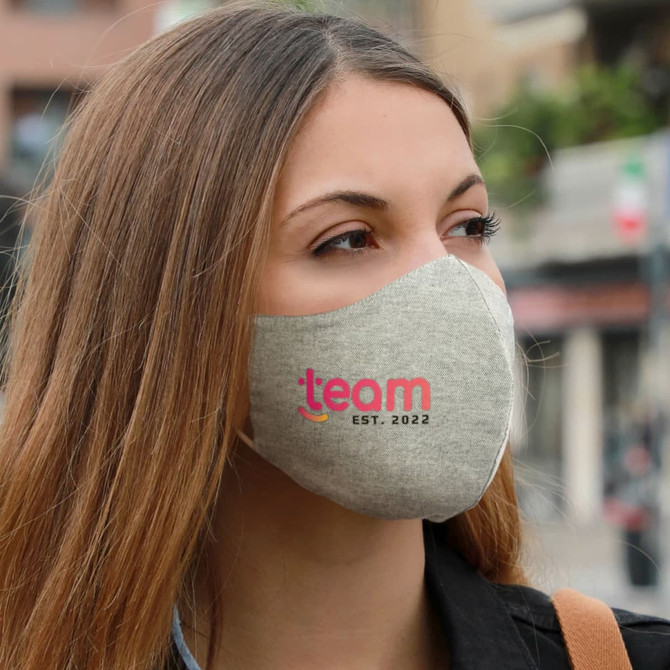 A linen face mask in light grey, featuring a logo in pink. The mask is worn by a person outdoors.