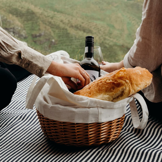 A wicker tote bag filled with a loaf of bread and a bottle of wine, set on a striped blanket outdoors.