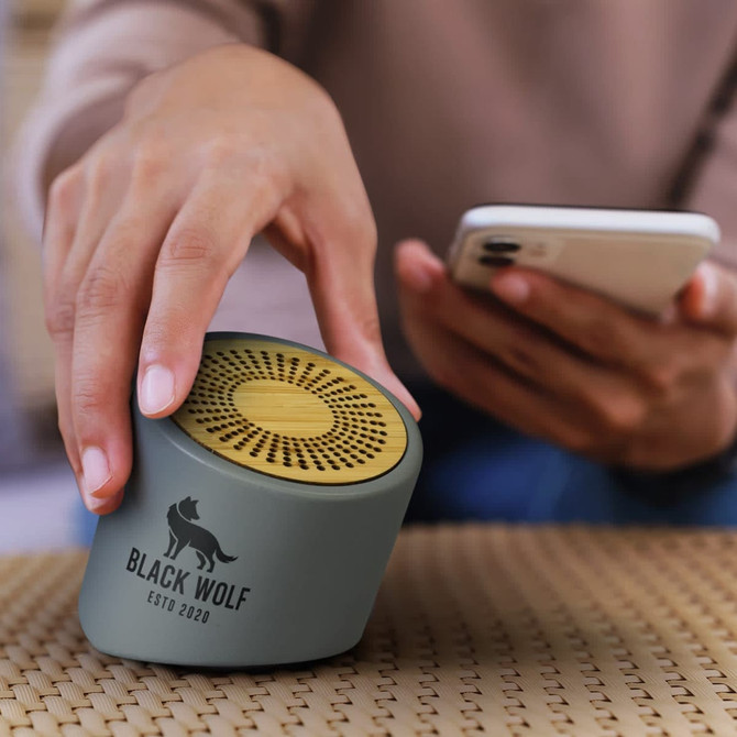 A green and gold NATURA Limestone Bluetooth mini speaker is being held next to a smartphone on a textured surface.