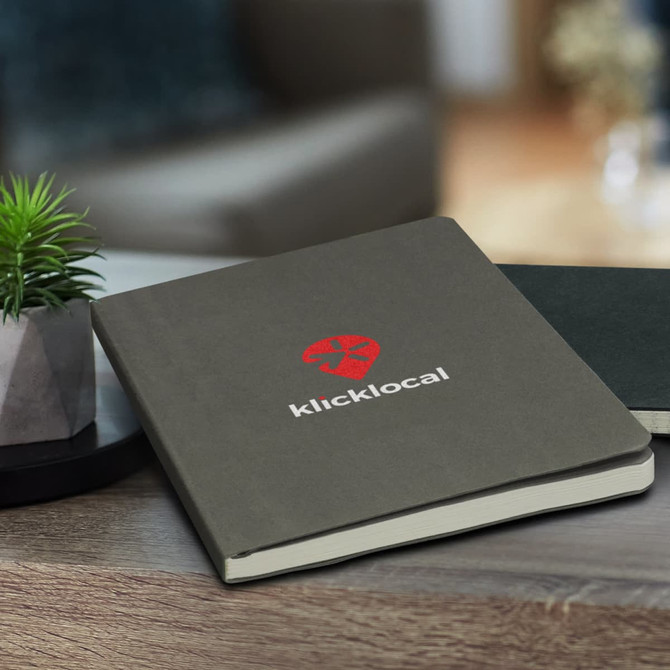 A grey recycled cotton soft cover notebook with a logo on the front, on a wooden surface next to a small plant.