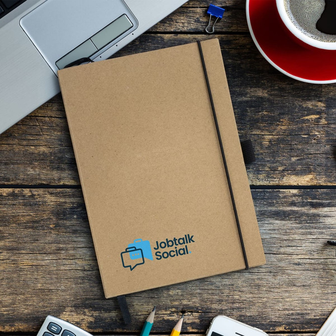 A brown hardcover notebook with an elastic band, featuring a logo, placed on a wooden desk alongside a coffee cup and stationery.