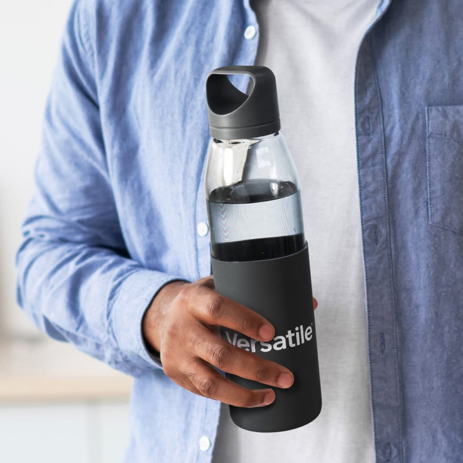 A drink bottle with a transparent glass body and a black silicone sleeve, held by a person wearing a blue shirt.