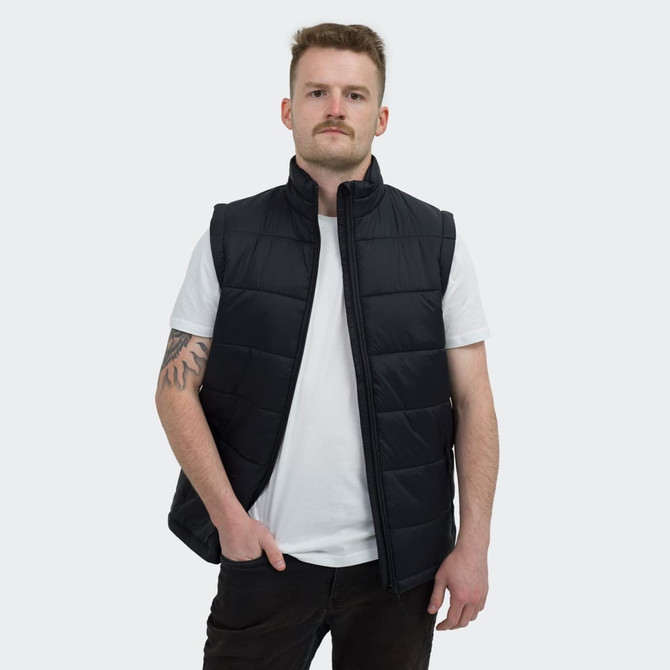 A man wearing a black puffer vest over a white t-shirt, standing with hands in pockets. The vest has a logo.