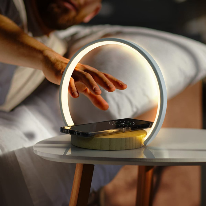 A white wireless charging night light with a circular design, illuminating a smartphone on a wooden table.