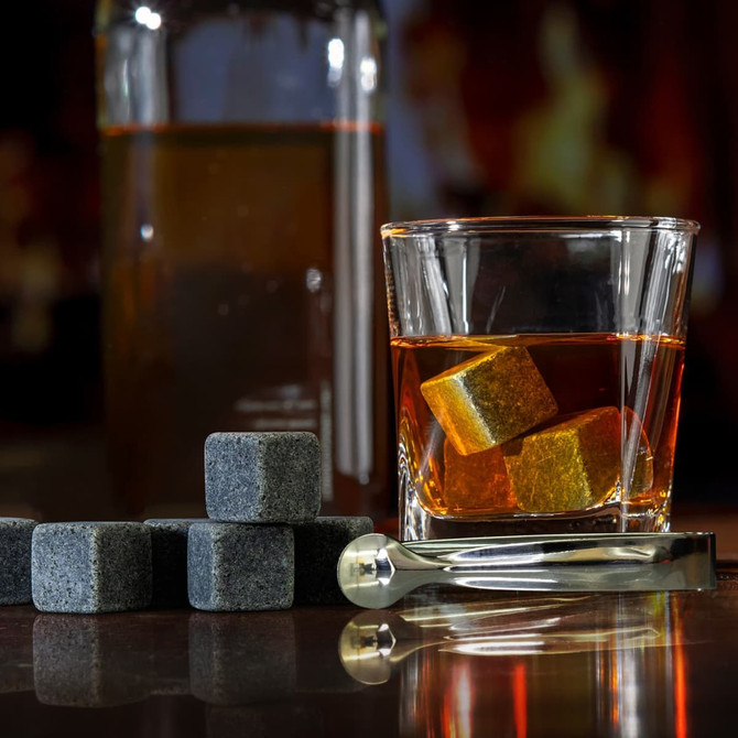 A glass with whiskey and grey whiskey stones beside a drink bottle and a stainless steel tongs.
