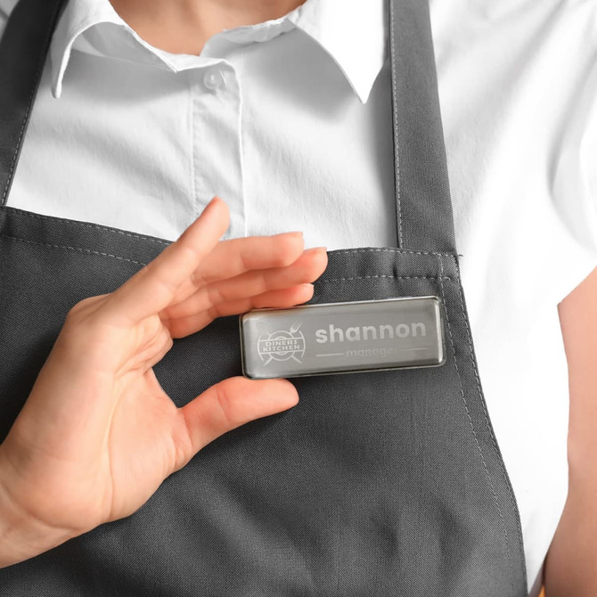 A metal name badge in silver is being held near a person's chest, showcasing its clear design and branding.