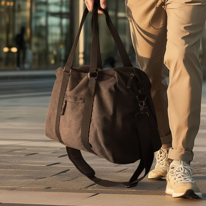 A dark brown canvas duffle bag with black straps, held by a person walking outdoors.