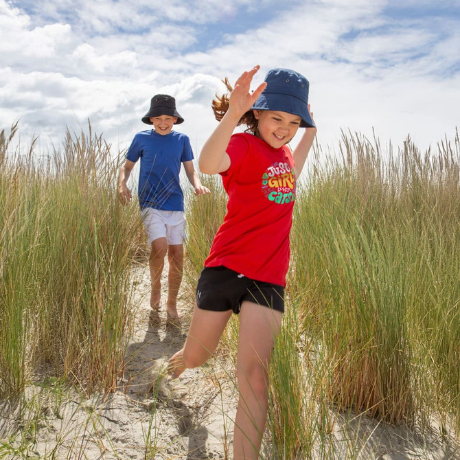 A red youth t-shirt worn by a girl running through tall grass, with a boy in a blue shirt nearby. Both wear hats.
