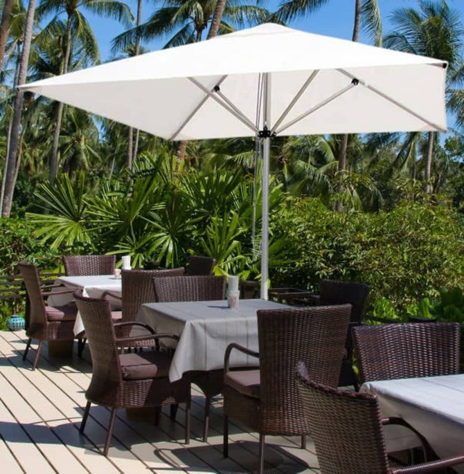 A white café umbrella with a sturdy frame stands over outdoor tables surrounded by lush greenery and palm trees.
