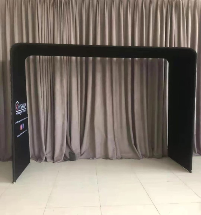 A black fabric arch frame with a tension design, set against a neutral curtain backdrop. Features a logo on one side.
