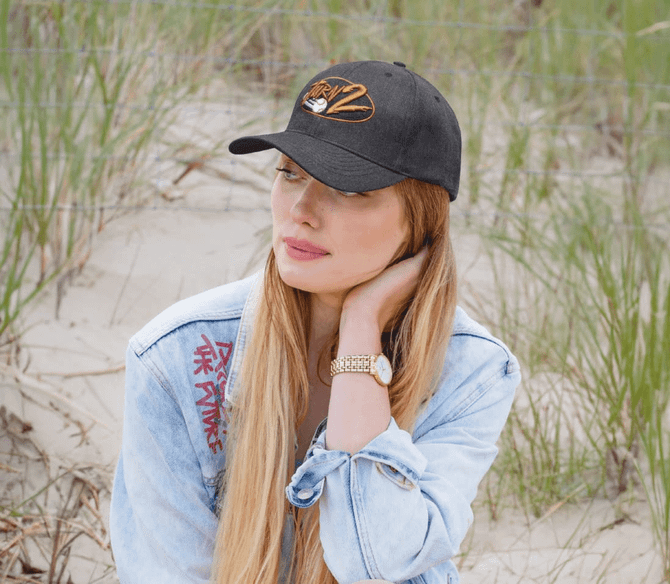 A black premium American twill cap with a logo, worn by a person with long hair, set against a sandy background.