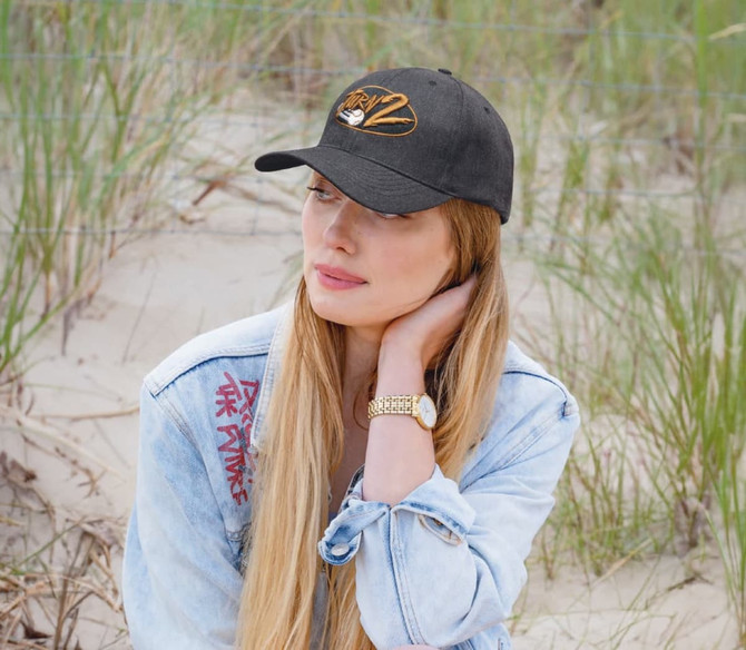 A woman with long blonde hair wears a black premium twill cap, seated on sandy ground with grass in the background.